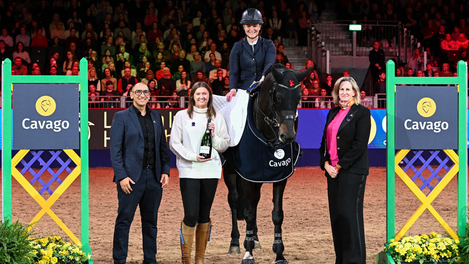 Equestrian rider on a horse with three people beside them in an indoor arena, with 'Cavago' branding and a seated audience in the background.